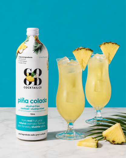 A bottle of COB Cocktail Co. Piña Colada cocktail and slushie mixer next to two tall glasses filled with a pale yellow drink and pineapple garnishes, set on a white marble surface against a bright turquoise background.